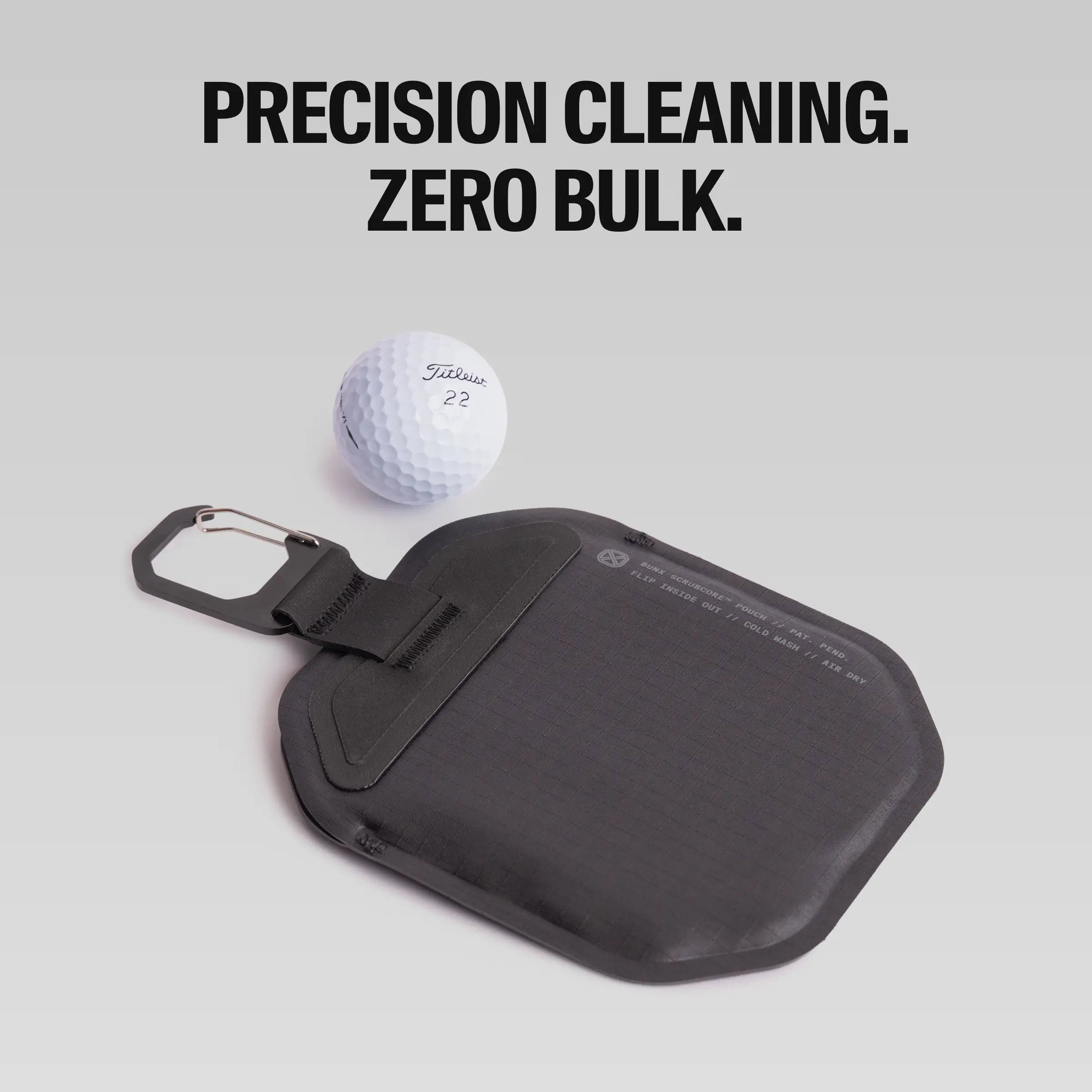 Black BUNX Golf Scrub Pouch lying flat beside a golf ball showing slim low-bulk profile